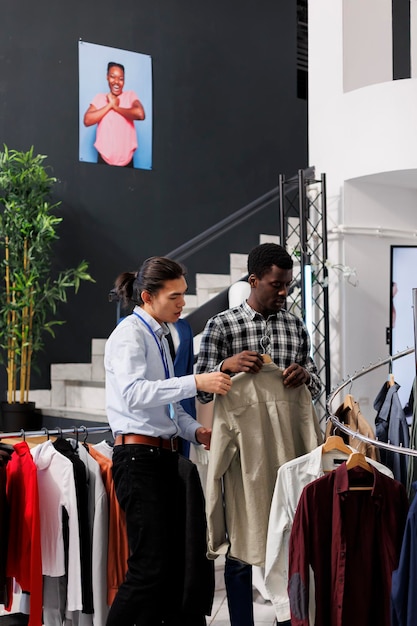 Un client afro-américain vérifie une chemise à la mode, discute du matériel vestimentaire avec un employé dans une boutique moderne. Un homme shopaholic fait du shopping pour des tenues à prix réduit, achète des vêtements décontractés. Concept de mode