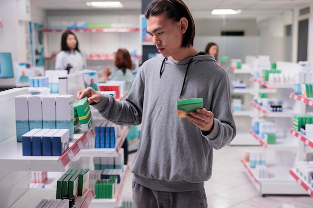 Client asiatique masculin examinant des suppléments sur des étagères, client regardant des boîtes de pilules dans une pharmacie remplie de produits de soins de santé comme des vitamines et des médicaments. Recherche de fournitures médicales.