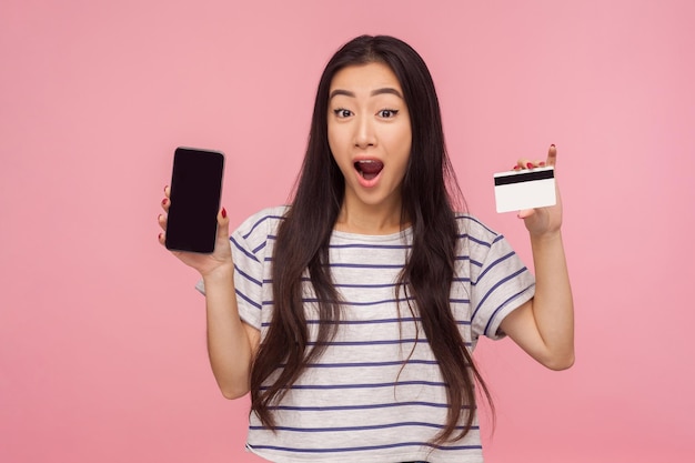 Client De Banque Choqué Par Une Bonne Banque Mobile. Portrait D'une Fille Surprise étonnée Aux Cheveux Bruns En T-shirt Rayé Tenant Une Carte De Crédit Et Un Téléphone Portable. Studio D'intérieur Tourné Isolé Sur Fond Rose