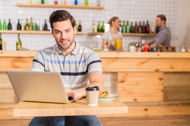 Client de café. Joyeux homme gentil positif assis à la table et travaillant sur l'ordinateur portable tout en déjeunant