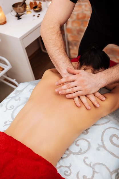 Client partiellement nu. Homme fort actif travaillant dans une armoire de massage avec une cliente aux cheveux noirs et réchauffant son dos