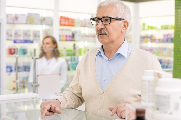 Client senior de pharmacie contemporaine debout au comptoir et attendant que le pharmacien le consulte sur le médicament à acheter