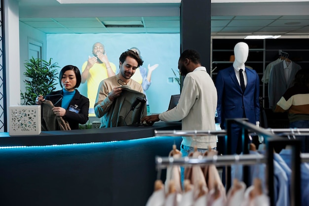 Client souriant et caissier discutant à la caisse tout en achetant des vêtements dans le département de mode du centre commercial. Un employé du magasin scanne un vêtement et discute avec un client afro-américain