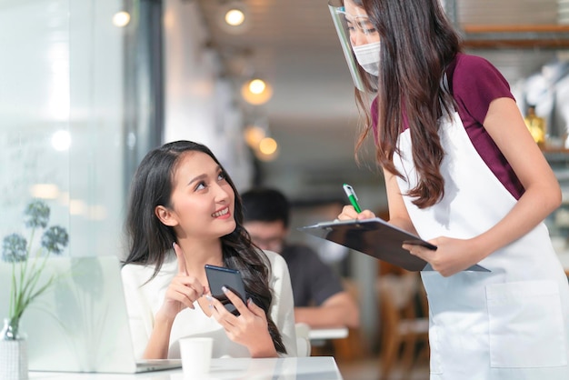 Une Cliente Asiatique Commande De La Nourriture à Partir D'un Menu Numérique De Smartphone Sans Contact Social Au Personnel De Serveuse Souriante Serveuse Du Millénaire Avec Carte Papier Prenant La Commande D'une Jeune Cliente Asiatique