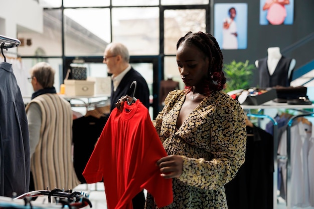 Une cliente enceinte achète des vêtements de grossesse élégants dans un magasin de vêtements, vérifie le tissu du t-shirt. Femme afro-américaine achetant des marchandises à la mode pour la maternité dans une salle d'exposition moderne