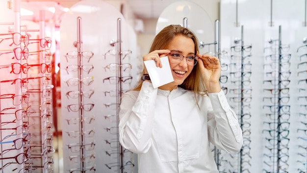 Une cliente ou un opticien heureux se tient debout avec des lunettes brutes en arrière-plan dans un magasin d'optique Stand avec des lunettes Correction de la vue Une fille à lunettes pose devant la caméra