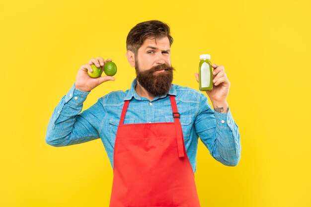 Clignant de l'homme en tablier tenant des citrons verts et une bouteille de jus fond jaune barman de jus