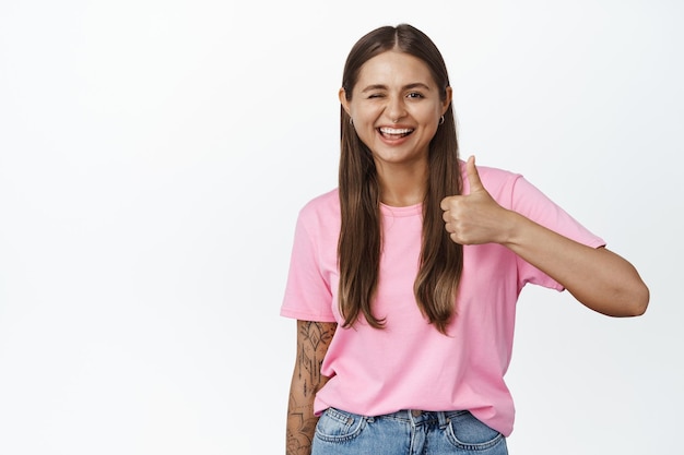 Clignotant fille souriante et montrant les pouces vers le haut, portant un t-shirt rose, debout sur fond blanc.