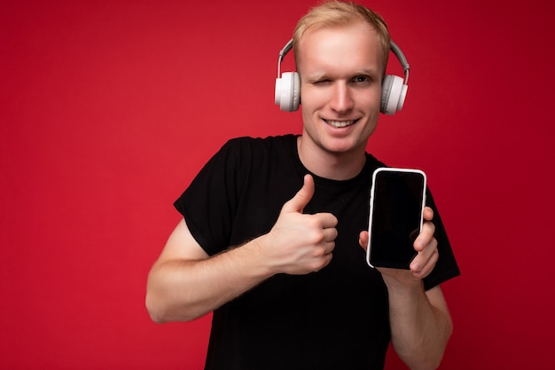 Un clin de œil beau jeune homme blond portant un t-shirt noir et un casque blanc isolé debout