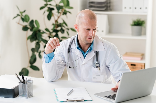 Clinicien chauve en uniforme à la recherche d'informations en ligne alors qu'il était assis par 24 devant un ordinateur portable