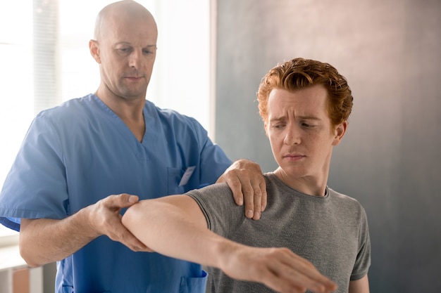 Clinicien de sexe masculin mature en uniforme bleu soutenant le bras courbé d'un jeune patient pendant la formation en physiothérapie dans des cliniques de réadaptation modernes