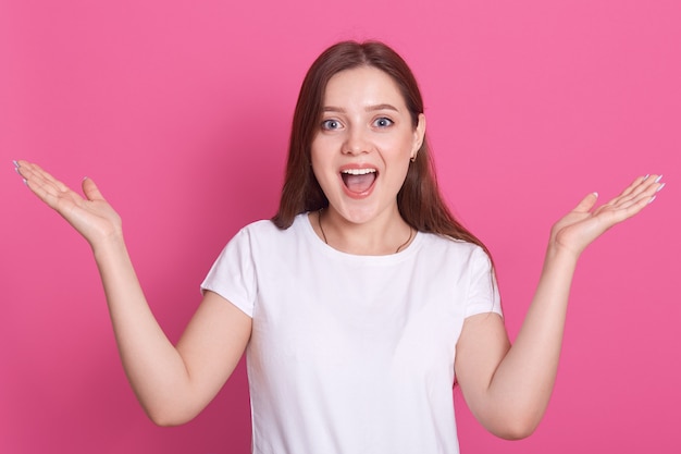 Clopse up amusé jeune femme portant un t-shirt décontracté blanc, tenant les mains levées, regardant directement la caméra et criant quelque chose. rose.