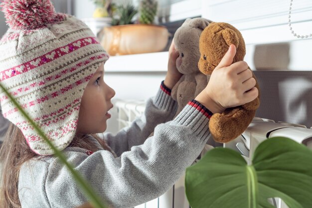 Close up calme petite fille concentrée en pull bonnet tricoté se réchauffer avec les meilleurs amis jouets en peluche près du radiateur de chaleur