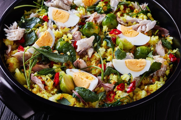 Close-up de délicieux Kedgeree avec du poisson fumé en flocons, des œufs durs, du riz, du chou frisé, des choux de Bruxelles, des épices et des herbes dans un four hollandais sur une table en bois noir, vue de dessus,