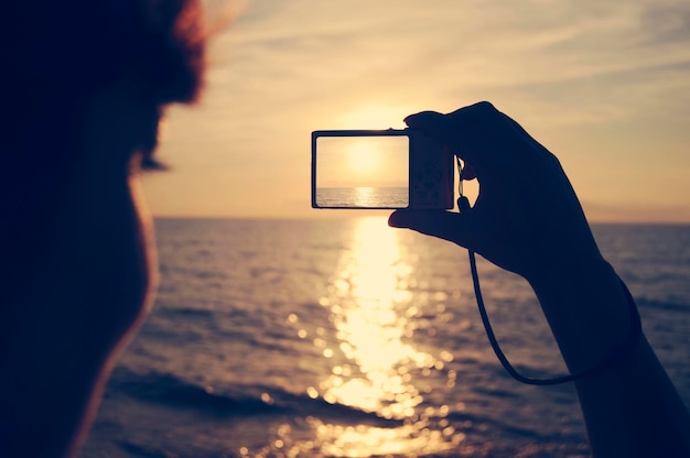 Photo close-up d'une femme photographiant la mer avec un appareil photo numérique contre le ciel au coucher du soleil