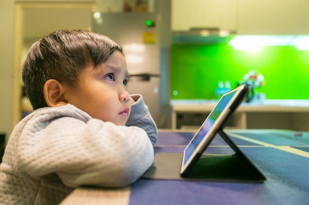 Close-up d'un garçon qui détourne son regard alors qu'il est assis avec une tablette numérique à la maison