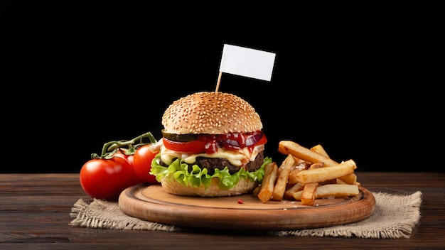 Close-up de hamburger fait maison avec du bœuf, des tomates, de la laitue, du fromage et des frites sur une planche à découper. Petit drapeau blanc inséré dans le burger