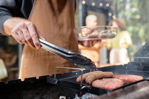 Close-up d'un jeune homme en tablier faisant des saucisses sur le gril pendant un pique-nique