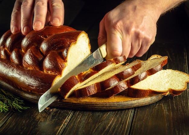 Close-up des mains d'un cuisinier coupées avec un couteau pain frais ou kalach sur une planche à couper de cuisine pour le petit déjeuner Nourriture saine et concept de boulangerie traditionnelle