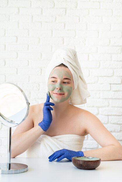 Close Up Of Happy Young Woman In White Serviettes De Bain Portant Des Gants Appliquant Un Masque D'argile En Regardant Le Miroir