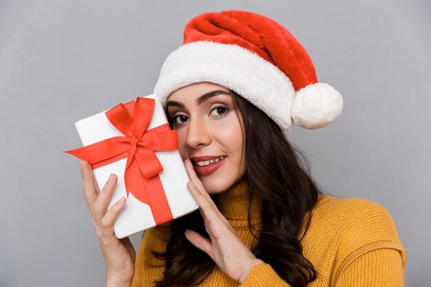 Close Up Of Smiling Young Woman Wearing Christmas Hat Debout Isolé Sur Fond Gris, Tenant Présent Fort