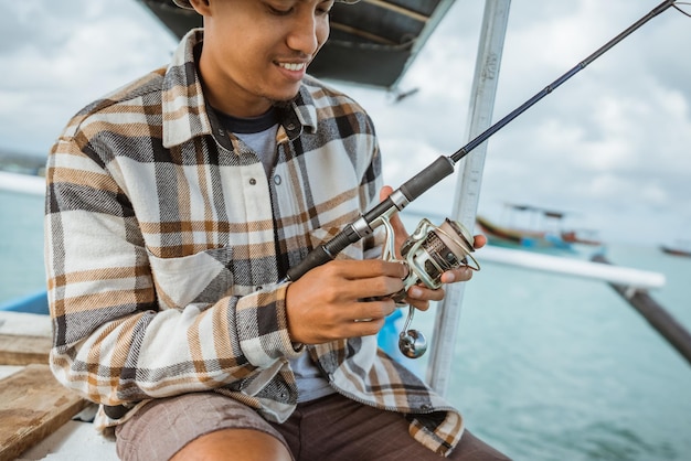 Close-up d'un pêcheur masculin asiatique vérifiant la bobine tout en tenant une canne à pêche sur un petit bateau de pêche