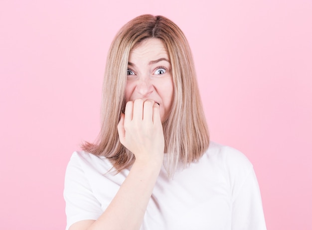 Close up portrait d'une adolescente inquiète en t-shirt blanc se mordant les ongles isolés sur rose