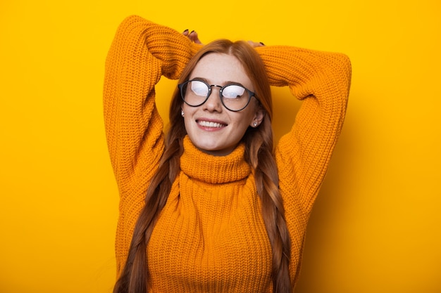 Close up portrait of ginger caucasian lady avec des taches de rousseur portant des lunettes et posant avec les mains sur un mur jaune