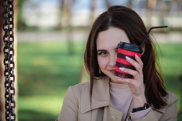 Close-up Portrait Of A Girl, Une Partie De Son Visage Recouvert D'un Verre Avec Une Paille. Verre Jetable Avec Une Boisson Dans Des Mains Féminines.