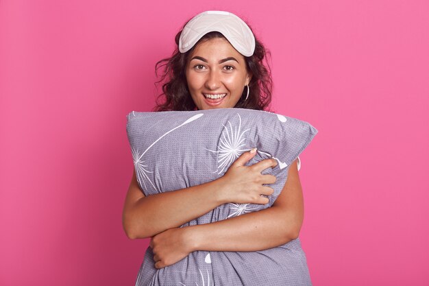 Close up portrait of happy young woman in pyjama and eye sleep mask hugging pillow over pink