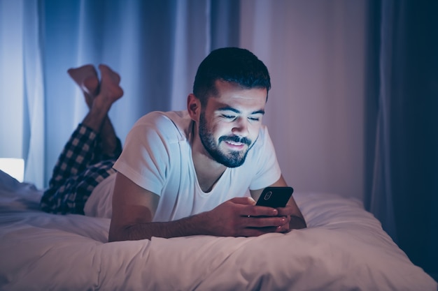 Close-up portrait of his he nice attractive gaie joyeuse brunette guy allongé sur le lit à l'aide du service de date de cellule envoi de sms week-end de temps libre gratuit dans la nuit en fin de soirée maison chambre d'hôtel à l'intérieur