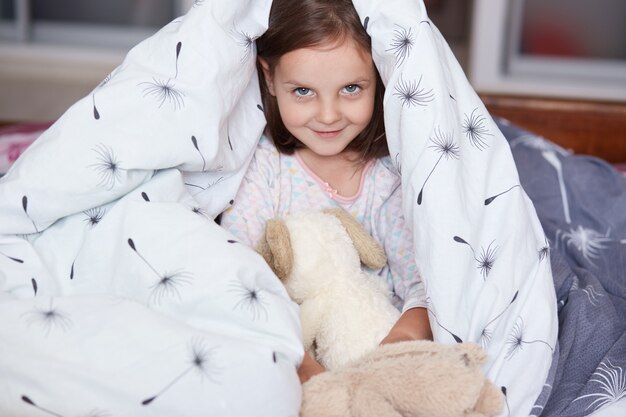 Photo close up portrait of little cheerful girl se cache sous une couverture avec jouet pour chien moelleux