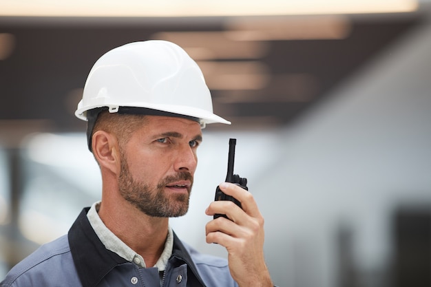 Close up portrait of mature worker parlant par talkie-walkie tout en supervisant les travaux sur chantier ou en atelier industriel,