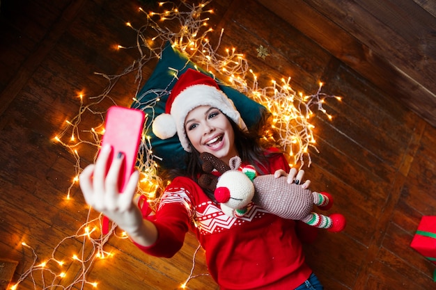 Close-up portrait of woman in Santa hat enveloppé dans des lumières de Noël prenant selfie et allongé sur le plancher en bois à la maison