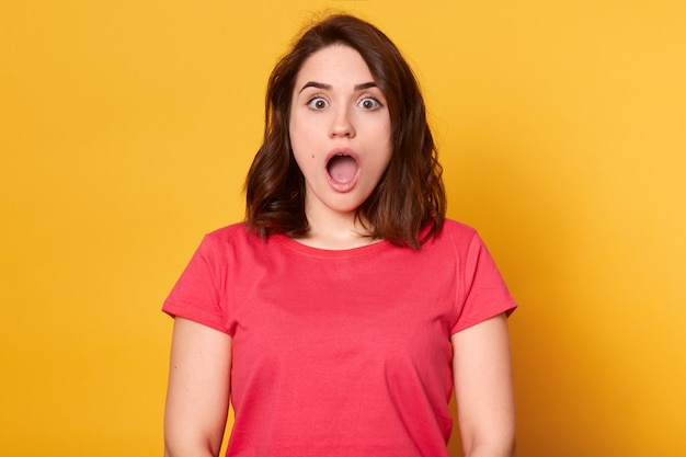 Close up portrait of young brunette woman looking surpris, debout avec la bouche grande ouverte, portant un t-shirt rouge isolé sur jaune