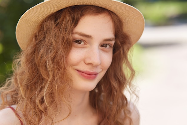 Close up portrait en plein air de jeune femme séduisante portant un chapeau de paille, a des cheveux ondulés foxy, regardant directement, a un sourire charmant