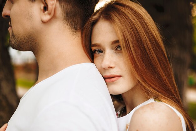 Close up portrait si une jolie femme aux cheveux rouges avec des taches de rousseur à la recherche directement sérieuse tout en penchant sa tête sur son petit ami à l'extérieur.