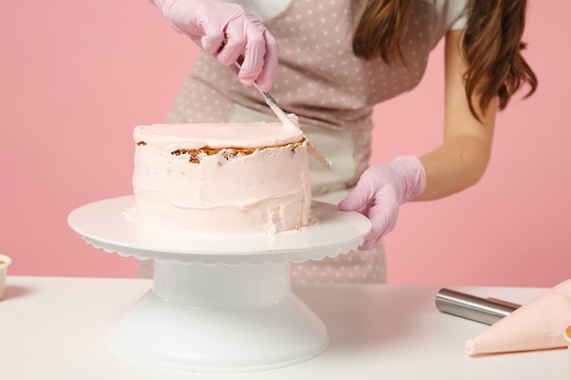 Close up recadrée chef cuisinier confiseur ou boulanger en t-shirt blanc cuisine à table isolé sur fond pastel rose en studio. Processus de fabrication de gâteau d'application de crème. Maquette du concept de nourriture de l'espace de copie.