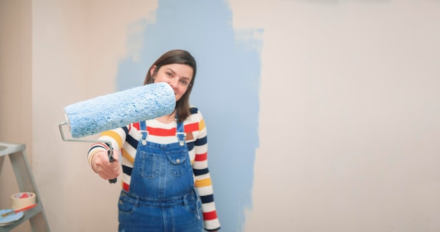 Close-up d'un rouleau avec de la peinture bleue dans la main d'une femme debout vêtue d'une combinaison