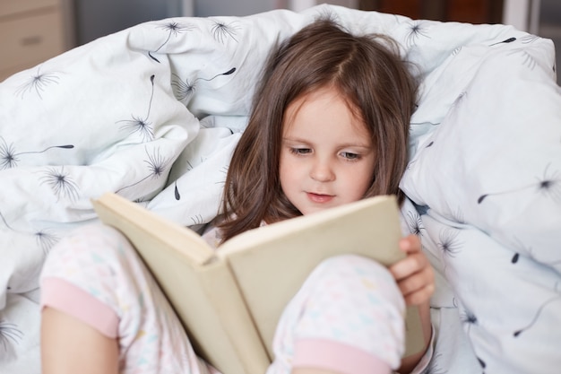 Close up shot intérieur de petite fille mignonne robes pyjama lire un livre intéressant en position couchée dans son lit