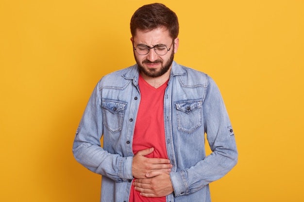 Close up studio portrait of bouleversé triste gars inquiet ayant des maux d'estomac, homme troublé portant une veste en jean et un t-shirt décontracté rouge, posant isolé sur jaune