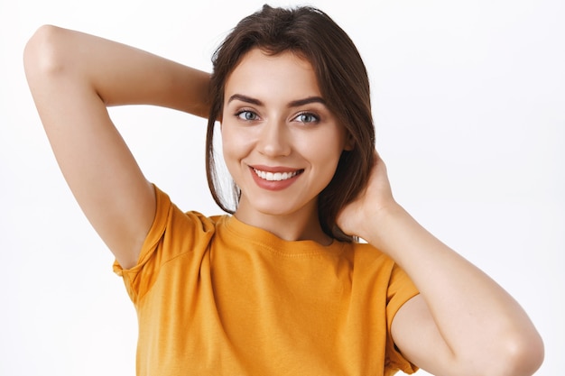 Close-up tendre, féminine séduisante jeune femme caucasienne élégante en t-shirt jaune, tenant les mains derrière pour réparer ses cheveux, faire une tresse ou le brosser, souriant heureux, fond blanc