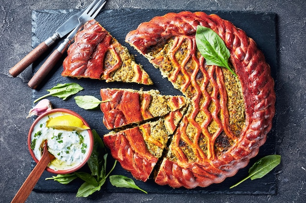 Close-up de tranches de tarte aux épinards Ricotta sur un plateau en ardoise noire avec sauce tzatziki dans un bol sur une table en béton, vue d'en haut, mise à plat