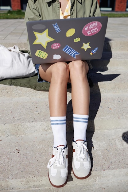 Close-up vertical d'un jeune étudiant méconnaissable utilisant un ordinateur portable à l'extérieur assis sur des escaliers en ville en se concentrant sur des jambes féminines portant des chaussettes