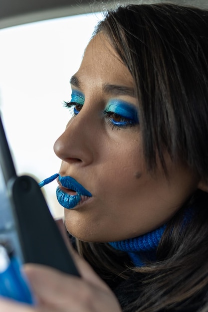 Close up vertical d'une jeune femme faisant son maquillage à l'intérieur d'une voiture avec des tons bleus regardant dans un miroir