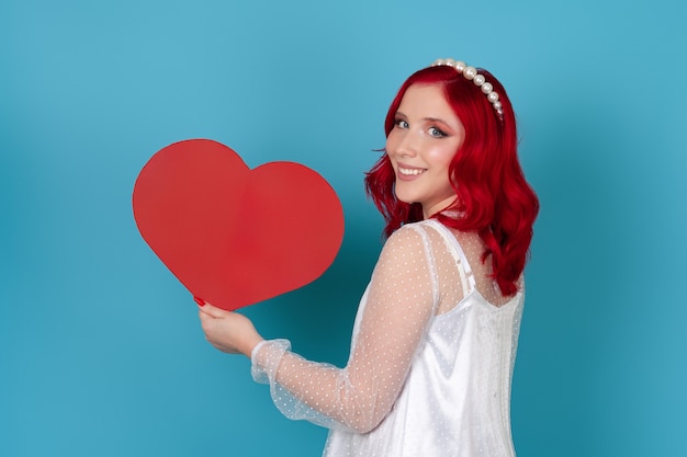 Close-up woman se tient de profil dans une robe blanche tenant un grand coeur de papier rouge