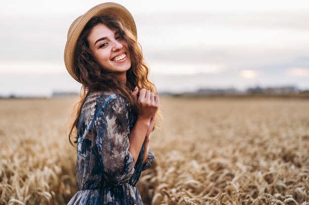 Closeup portrait d'une belle jeune femme aux cheveux bouclés. Femme en robe et chapeau debout dans le champ de blé