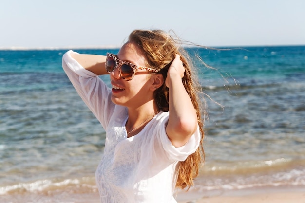 Closeup portrait d'une belle jeune fille brune aux cheveux longs sur un fond de mer bleue avec des vagues et du ciel avec des nuages sur un mode de vie ensoleillé posant et souriant vent