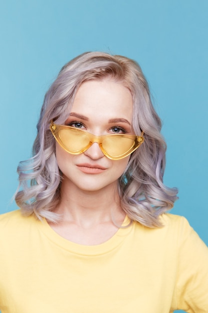 Closeup portrait de femme blonde dans les vêtements jaunes et lunettes de soleil isolées sur fond bleu.