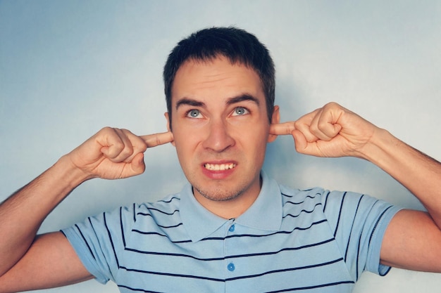 Closeup portrait jeune homme stressé malheureux en colère couvrant ses oreilles levant les yeux pour dire arrêter de faire du bruit fort, il donne des maux de tête fond bleu Émotions négatives expressions du visage